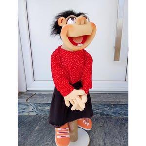 May include: A handmade puppet with a smiling face, black hair, and a red polka-dot shirt. The puppet wears a black skirt and orange sneakers. The puppet's hands are clasped together. The puppet is standing on a wooden stand.