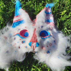 Può includere: Una maschera da animale in pelliccia sintetica bianca con orecchie a punta decorate da strisce blu e rosa. La maschera presenta orbite oculari rosa con centri blu, un ponte nasale rosa e un naso blu brillante. La pelliccia bianca è decorata con piccole macchie rosa e blu.