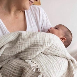 Op de afbeelding: Een vrouw die een baby vasthoudt die gewikkeld is in een zachte, witte muslin-wikkeldeken met franjes. De deken is gemaakt van een lichtgewicht, ademende stof.
