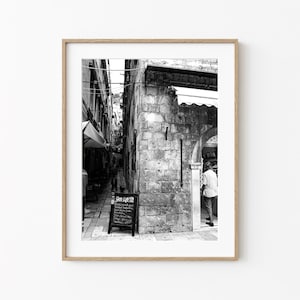 Puede incluir: Impresión enmarcada en blanco y negro de una escena de calle europea. Un edificio de piedra con una puerta arqueada está a la derecha. Una pizarra está frente a un edificio a la izquierda. Una persona camina por la puerta.