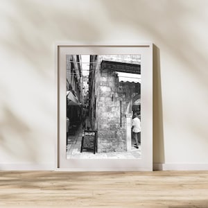 May include: Black and white framed photograph of a narrow European street scene. The image features stone buildings, an arched doorway, and a chalkboard sign. A person is seen walking through the doorway.