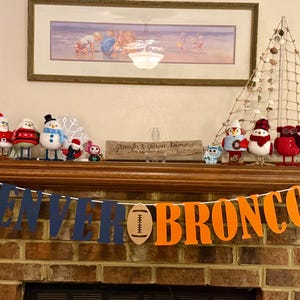 May include: A decorated mantelpiece with a banner that reads "DENVER BRONCOS" in navy blue and orange. The mantel is adorned with various Christmas-themed figurines, including snowmen, penguins, and owls. A framed picture hangs above.
