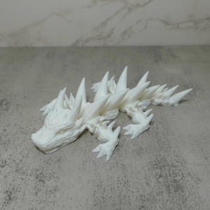 May include: A white, 3D printed model of a spiky, reptilian creature. The creature has a long, pointed tail and a wide, open mouth with sharp teeth.