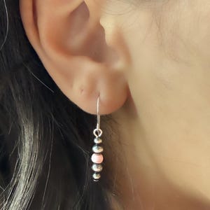 May include: A silver earring with a hook, featuring a series of small, round beads. The beads alternate between silver and pale pink, creating a delicate and feminine design. The earring is shown worn on an ear.