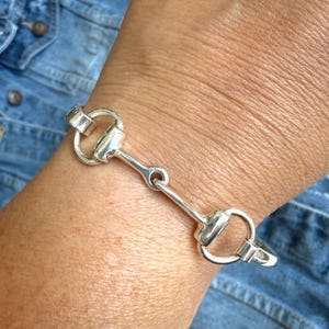 Könnte beinhalten: Ein poliertes Armband in Silberoptik mit einem zentralen Trensen-Design. Die Gliederkonstruktion besteht aus zwei kreisförmigen Ringen, die durch einen horizontalen Steg verbunden sind, was eine klassische, vom Reitsport inspirierte Optik mit glatter Oberfläche ergibt.
