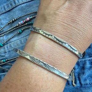 May include: Two silver cuff bracelets featuring a stamped sunburst pattern along the outer edge. The bracelets have a polished metallic finish and a thin, flat band design. The metal surface displays repeating geometric rays etched into the silver.
