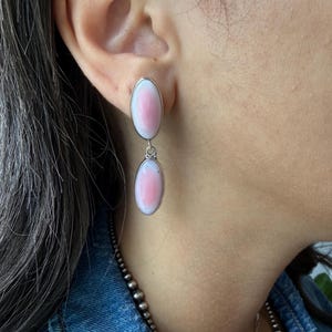 May include: A pair of drop earrings featuring two oval-shaped, smooth pink and white stones set in polished silver bezels. The earrings consist of a top oval stone attached to the earlobe and a second matching oval stone dangling below it.