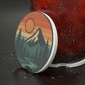 May include: An oval car coaster with a mountain and forest design. The coaster features a sunset with orange, yellow, and tan stripes. The mountains and trees are dark green and the coaster has a white border.