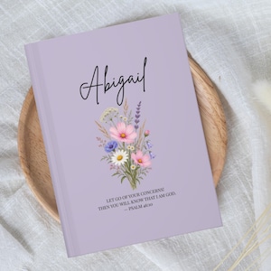 Puede incluir: Un cuaderno de color morado claro con el nombre Abigail en letra cursiva negra, una ilustración de un ramo de flores coloridas y el texto: LET GO OF YOUR CONCERNS! THEN YOU WILL KNOW THAT I AM GOD. — PSALM 46:10 impreso en negro.