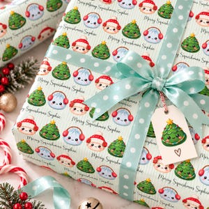 May include: Wrapping paper with a repeating pattern of cartoon Christmas trees, dumplings wearing red earmuffs, and the text Merry Squishmas Sophie. The gift is tied with a light blue ribbon featuring white polka dots and a gift tag.