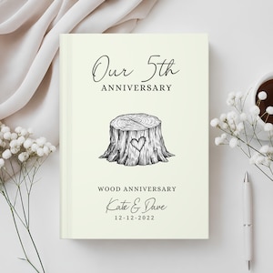 May include: A cream-coloured hardcover book with black text reading Our 5th ANNIVERSARY, WOOD ANNIVERSARY, Kate & Dave, and 12-12-2022. A black and white line drawing of a tree stump with a heart carved into the side is centred on the cover.