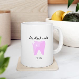 Puede incluir: Taza de cerámica blanca con asa, con el texto "Dr. Richards" encima de un gráfico de diente rosa con una cruz y el texto "EST 2026". La taza está sobre un posavasos beige.