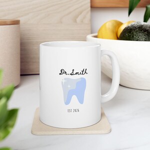 May include: White ceramic mug with a dental theme. The mug features the text "Dr. Smith" in black script above a light blue tooth graphic with a sparkle and the text "EST 2026" below the tooth. The mug sits on a beige coaster.