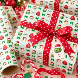 May include: Wrapping paper with a repeating pattern of green and red Christmas ornaments wearing Santa hats, green bows, and the name Sophie in script. The gift is tied with a red ribbon featuring white polka dots and a matching gift tag.