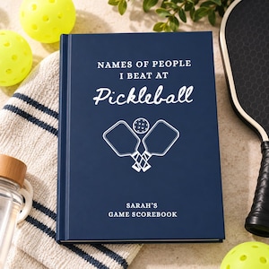 以下が含まれることがあります： NAMES OF PEOPLE I BEAT AT PickleballおよびSARAH'S GAME SCOREBOOKという白い文字が書かれたネイビーブルーのハードカバーブック。表紙の中央には、2本の交差したピックルボール用パドルとボールの白いグラフィックが描かれています。