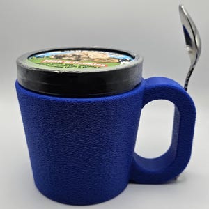 May include: A blue textured mug with a handle, holding a pint of ice cream. A silver spoon rests on the side. The ice cream container has a colorful label. The mug appears to be made of a 3D printed material.