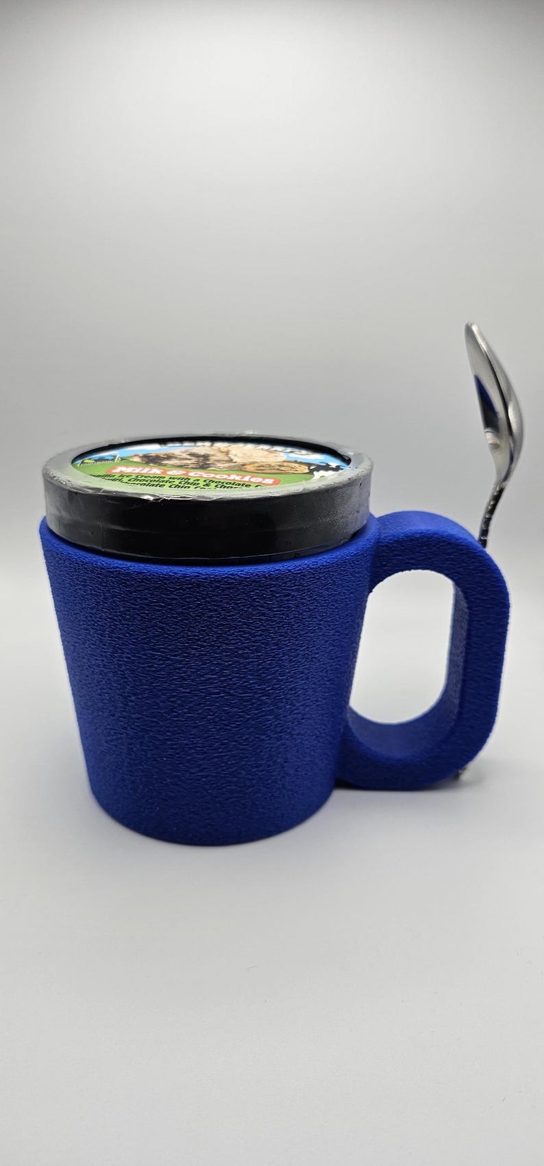 May include: A blue textured mug with a handle, holding a pint of ice cream. A silver spoon rests on the side. The ice cream container has a colorful label. The mug appears to be made of a 3D printed material.