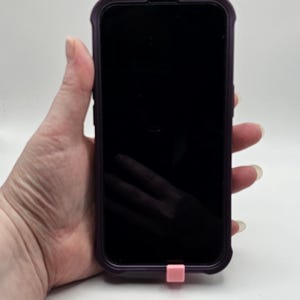 以下が含まれることがあります： 紫色のケースに入った黒いスマートフォンと、ピンク色の充電ポートカバー。スマートフォンの画面は黒です。白い背景に、スマートフォンが手に持たれています。