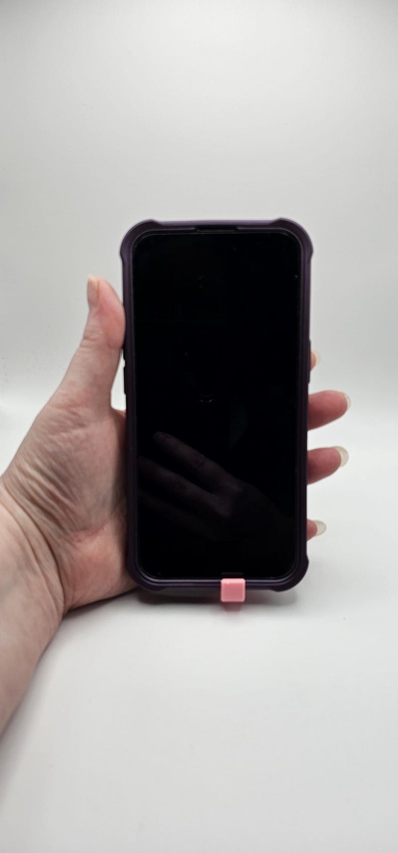 以下が含まれることがあります： 紫色のケースに入った黒いスマートフォンと、ピンク色の充電ポートカバー。スマートフォンの画面は黒です。白い背景に、スマートフォンが手に持たれています。