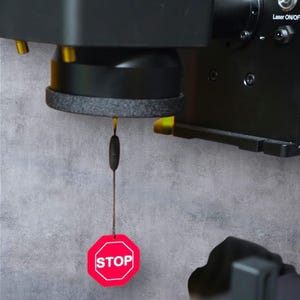 May include: A black and gray machine component with a red stop sign attached. The stop sign is octagonal with white text that reads "STOP". The machine has a black base and a gray textured ring.