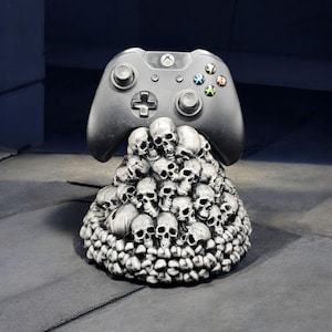 May include: A black Xbox controller rests atop a pile of detailed, gray skulls. The controller features colored buttons and a directional pad. The base of skulls is textured and appears to be made of stone, creating a unique gaming accessory.