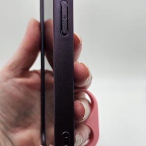 以下が含まれることがあります： 透明なスクリーンプロテクターが付いた、プラム色の携帯電話ケース。ケースは滑らかな仕上がりで、側面に音量ボタンと電源ボタンが付いています。ピンク色のループがケースに取り付けられています。