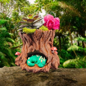 May include: A brown tree stump planter with a sleeping green dragon inside. The planter is decorated with a succulent, pink flowers, and small red mushrooms. The planter is set against a blurred green background.