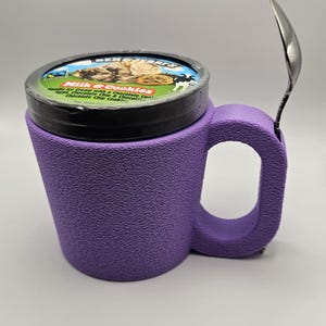 May include: A purple ice cream mug with a textured surface, holding a pint of ice cream. A silver spoon rests on the side. The ice cream container has a black lid with the brand name and flavor description: "Milk & Cookies".