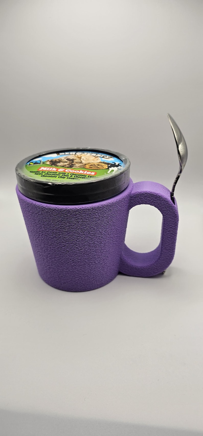 May include: A purple ice cream mug with a textured surface, holding a pint of ice cream. A silver spoon rests on the side. The ice cream container has a black lid with the brand name and flavor description: "Milk & Cookies".