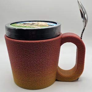 May include: A textured, gradient-colored mug with a lid and a spoon. The mug transitions from a reddish-brown to a yellow-orange hue. A small spoon rests on the side. The lid has a colorful image. The mug is designed for beverages.