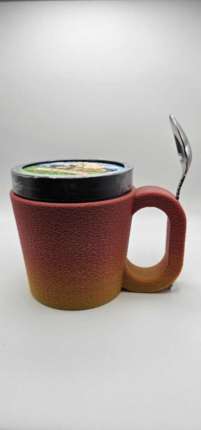May include: A textured, gradient-colored mug with a lid and a spoon. The mug transitions from a reddish-brown to a yellow-orange hue. A small spoon rests on the side. The lid has a colorful image. The mug is designed for beverages.