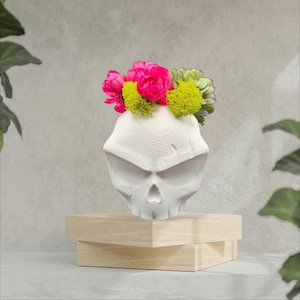May include: A white skull-shaped vase holding a vibrant arrangement of pink peonies, green moss, and succulents. The vase sits on a light-colored wooden stand, creating a unique decorative piece.