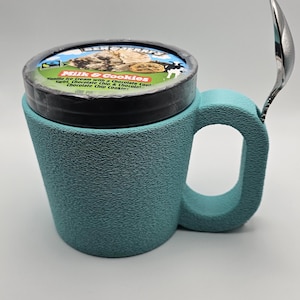 May include: A teal ice cream mug with a handle, holding a pint of ice cream. A spoon rests on the side. The ice cream container is labeled "Milk & Cookies" with a description of the flavors.