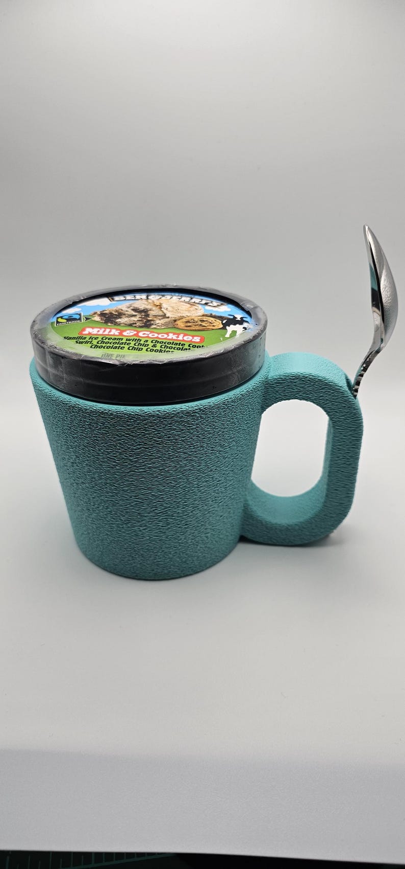 May include: A teal ice cream mug with a handle, holding a pint of ice cream. A spoon rests on the side. The ice cream container is labeled "Milk & Cookies" with a description of the flavors.