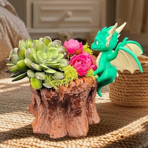 May include: A decorative planter shaped like a tree stump, filled with green succulents, pink flowers, and moss. A green dragon figurine with cream-colored wings is perched on the edge of the planter. The planter is brown.