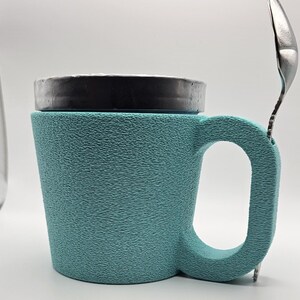 May include: A teal textured mug with a black lid and a silver spoon resting on the side. The mug has a large handle and a textured surface. The spoon is made of stainless steel.