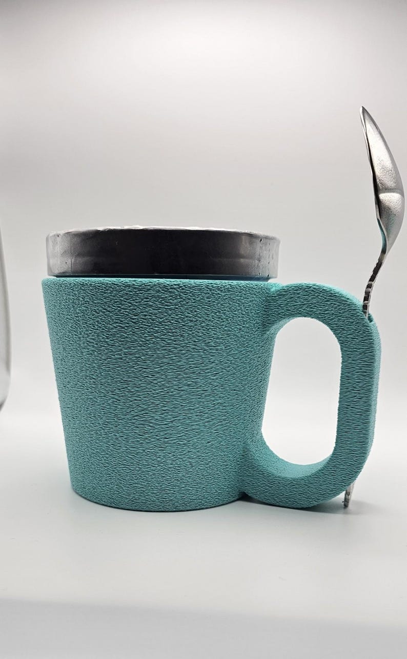 May include: A teal textured mug with a black lid and a silver spoon resting on the side. The mug has a large handle and a textured surface. The spoon is made of stainless steel.