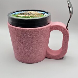 May include: A pink textured mug with a handle, holding a pint of ice cream with a black lid. A silver spoon rests on the side of the mug. The ice cream container has a label with text and images.