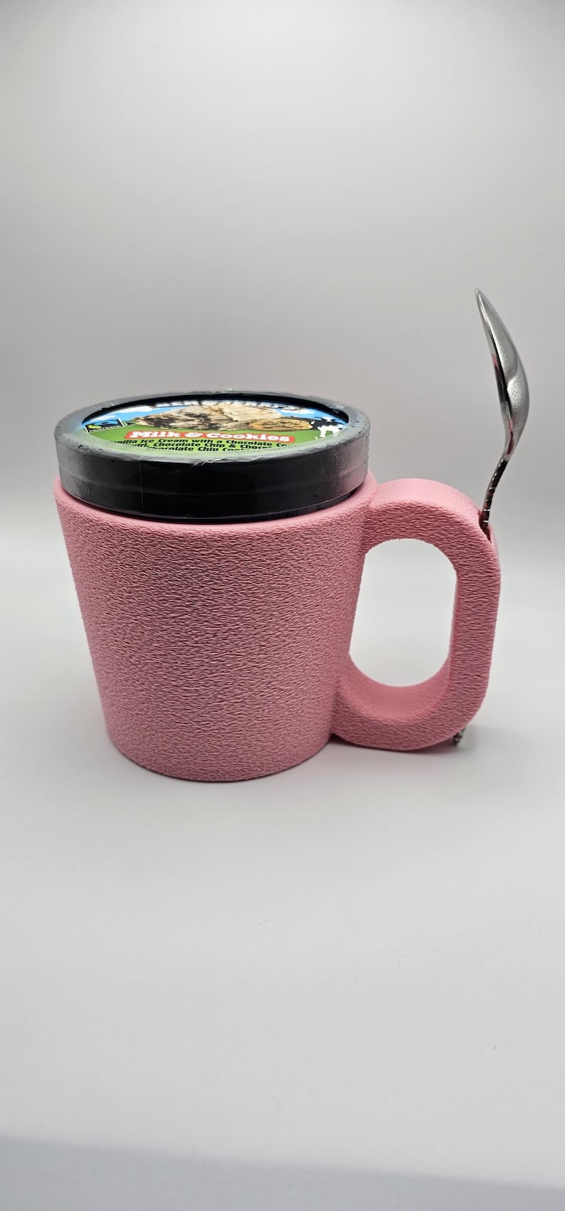 May include: A pink textured mug with a handle, holding a pint of ice cream with a black lid. A silver spoon rests on the side of the mug. The ice cream container has a label with text and images.