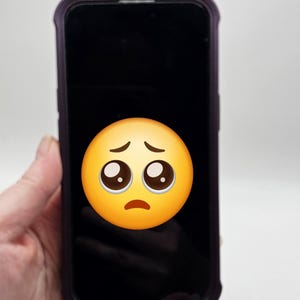 以下が含まれることがあります： 黒い画面と悲しい顔の絵文字が付いたスマートフォン。 電話は紫色のケースに入っており、「No Pinky Saver」と「ouch！」の文字が見えます。