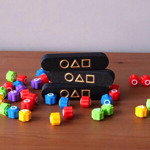 May include: A collection of colorful game pieces and three black rectangular blocks. The blocks are stacked and feature gold symbols. The game pieces are in various colors, including red, blue, green, and yellow. The game pieces are scattered on a wooden surface.