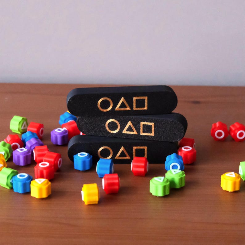 May include: A collection of colorful game pieces and three black rectangular blocks. The blocks are stacked and feature gold symbols. The game pieces are in various colors, including red, blue, green, and yellow. The game pieces are scattered on a wooden surface.