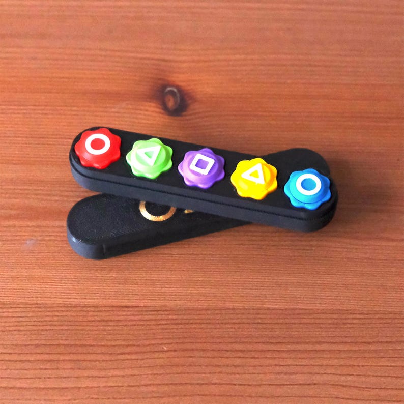 May include: Black controller-shaped accessory with five colorful buttons: red circle, green triangle, purple square, yellow triangle, and blue circle. The accessory is resting on a wooden surface.