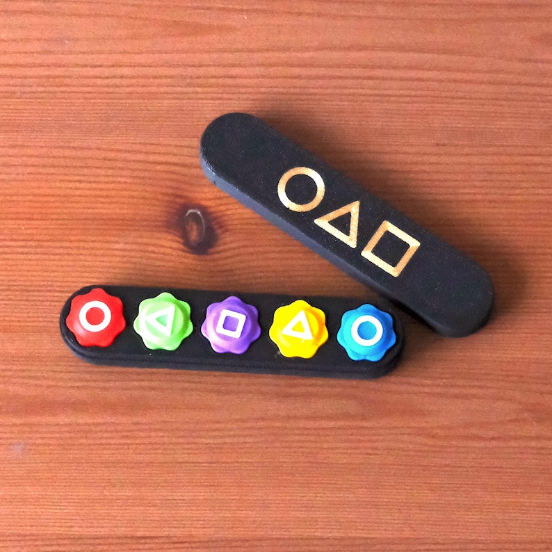 May include: Black game-themed accessories with gold lettering and colorful buttons. One piece has the text "O&Delta;□" in gold. The other piece has six colorful buttons with different shapes. The items are on a wooden surface.