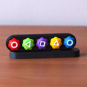 May include: A black plastic desk organizer with five colorful, flower-shaped buttons. Each button has a different shape in the center: a circle, triangle, square, triangle, and circle. The buttons are red, green, purple, yellow, and blue.