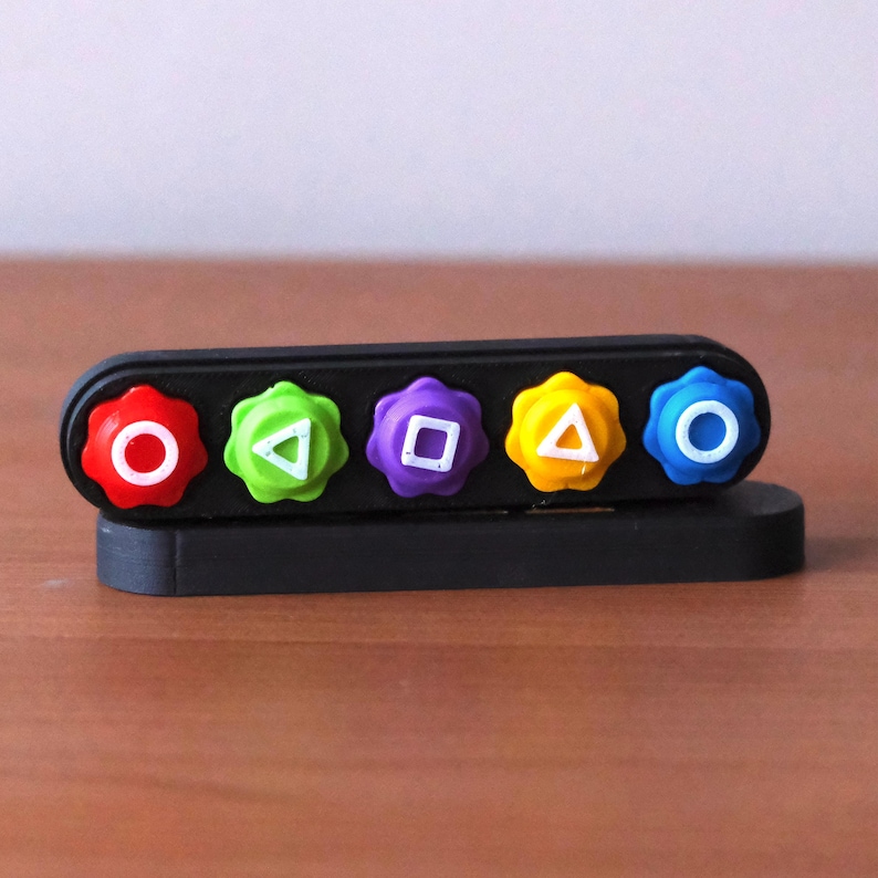 May include: A black plastic desk organizer with five colorful, flower-shaped buttons. Each button has a different shape in the center: a circle, triangle, square, triangle, and circle. The buttons are red, green, purple, yellow, and blue.