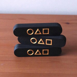 May include: Three black, rectangular wooden blocks stacked on top of each other. Each block has a gold symbol consisting of a circle, a triangle, and a square. The blocks are resting on a wooden surface.