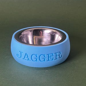 Könnte beinhalten: Eine hellblaue Futterschale für Haustiere mit einem Einsatz aus Edelstahl. Die Schale hat das Wort "JAGGER" auf der Seite eingeprägt.