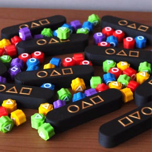 May include: A collection of black game pieces with gold symbols and colorful, geometric shapes. The shapes are red, yellow, green, blue, and purple. The game pieces are arranged on a wooden surface.