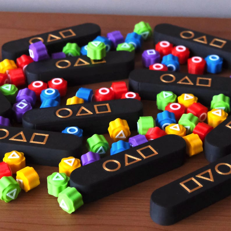 May include: A collection of black game pieces with gold symbols and colorful, geometric shapes. The shapes are red, yellow, green, blue, and purple. The game pieces are arranged on a wooden surface.